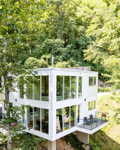 Spacious living room with vaulted wood ceiling, ceiling fan, brown leather furniture, fireplace, and expansive windows overlooking forested mountains and